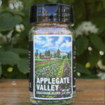 Glass jar of “Applegate Valley” seasoning blend sitting on a wooden table outdoors with blurred green leaves and white flowers in the background. The label features illustrated rows of crops on a farm beneath a blue sky.
