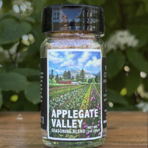 Glass jar of “Applegate Valley” seasoning blend sitting on a wooden table outdoors with blurred green leaves and white flowers in the background. The label features illustrated rows of crops on a farm beneath a blue sky.