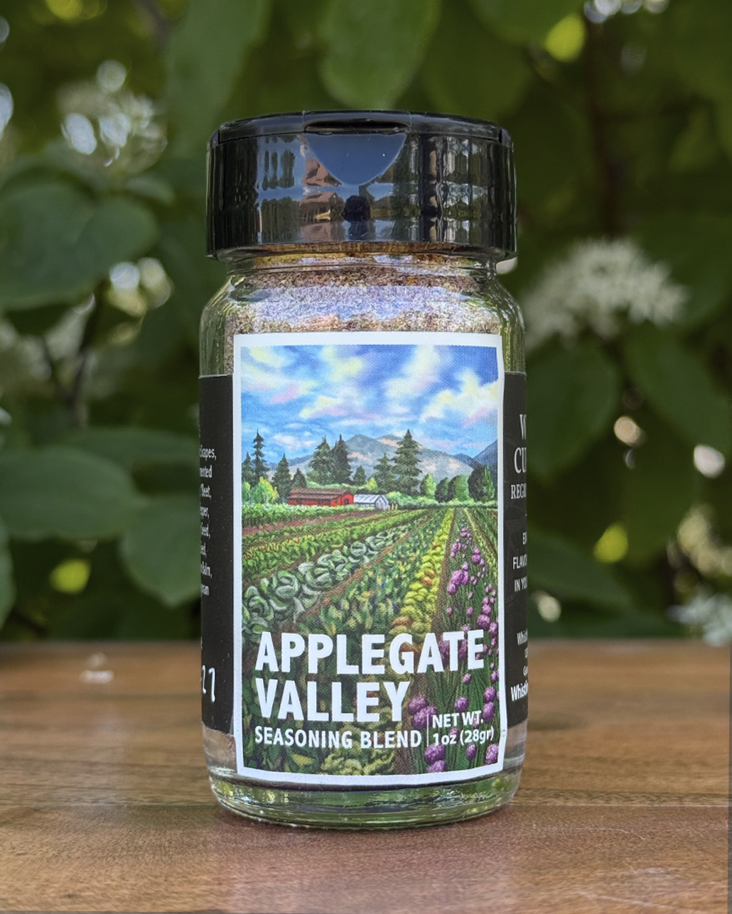 Glass jar of “Applegate Valley” seasoning blend sitting on a wooden table outdoors with blurred green leaves and white flowers in the background. The label features illustrated rows of crops on a farm beneath a blue sky.