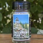 Glass jar of “Dutchman Peak” seasoning blend sitting on a wooden table outdoors with blurred green leaves and white flowers in the background. The label features an illustrated historic fire lookout on a mountaintop beneath a blue sky.