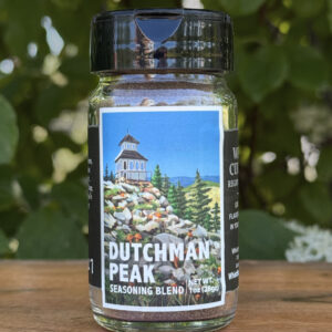 Glass jar of โDutchman Peakโ seasoning blend sitting on a wooden table outdoors with blurred green leaves and white flowers in the background. The label features an illustrated historic fire lookout on a mountaintop beneath a blue sky.