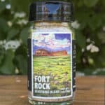 Glass jar of “Fort Rock” seasoning blend sitting on a wooden table outdoors with blurred green leaves and white flowers in the background. The label features an illustrated desert scape beneath a blue sky.