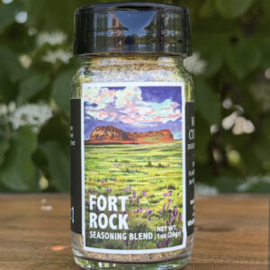 Glass jar of โFort Rockโ seasoning blend sitting on a wooden table outdoors with blurred green leaves and white flowers in the background. The label features an illustrated desert scape beneath a blue sky.