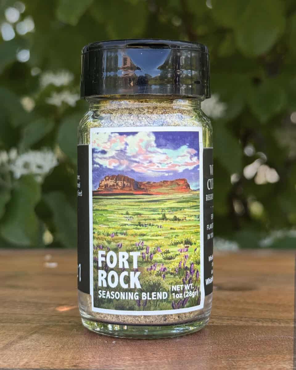 Glass jar of “Fort Rock” seasoning blend sitting on a wooden table outdoors with blurred green leaves and white flowers in the background. The label features an illustrated desert scape beneath a blue sky.