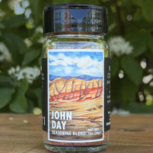 Glass jar of โJohn Dayโ seasoning blend sitting on a wooden table outdoors with blurred green leaves and white flowers in the background. The label features an illustrated desert scape beneath a blue sky.