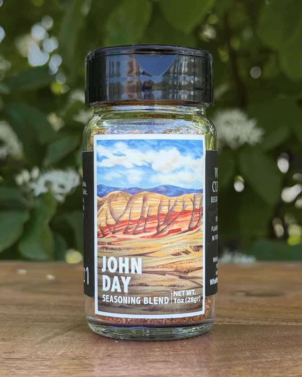 Glass jar of “John Day” seasoning blend sitting on a wooden table outdoors with blurred green leaves and white flowers in the background. The label features an illustrated desert scape beneath a blue sky.
