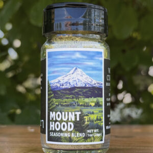 Glass jar of “Mount Hood” seasoning blend sitting on a wooden table outdoors with blurred green leaves and white flowers in the background. The label features an illustrated ice-capped mountain with a lush green foreground beneath a blue sky.