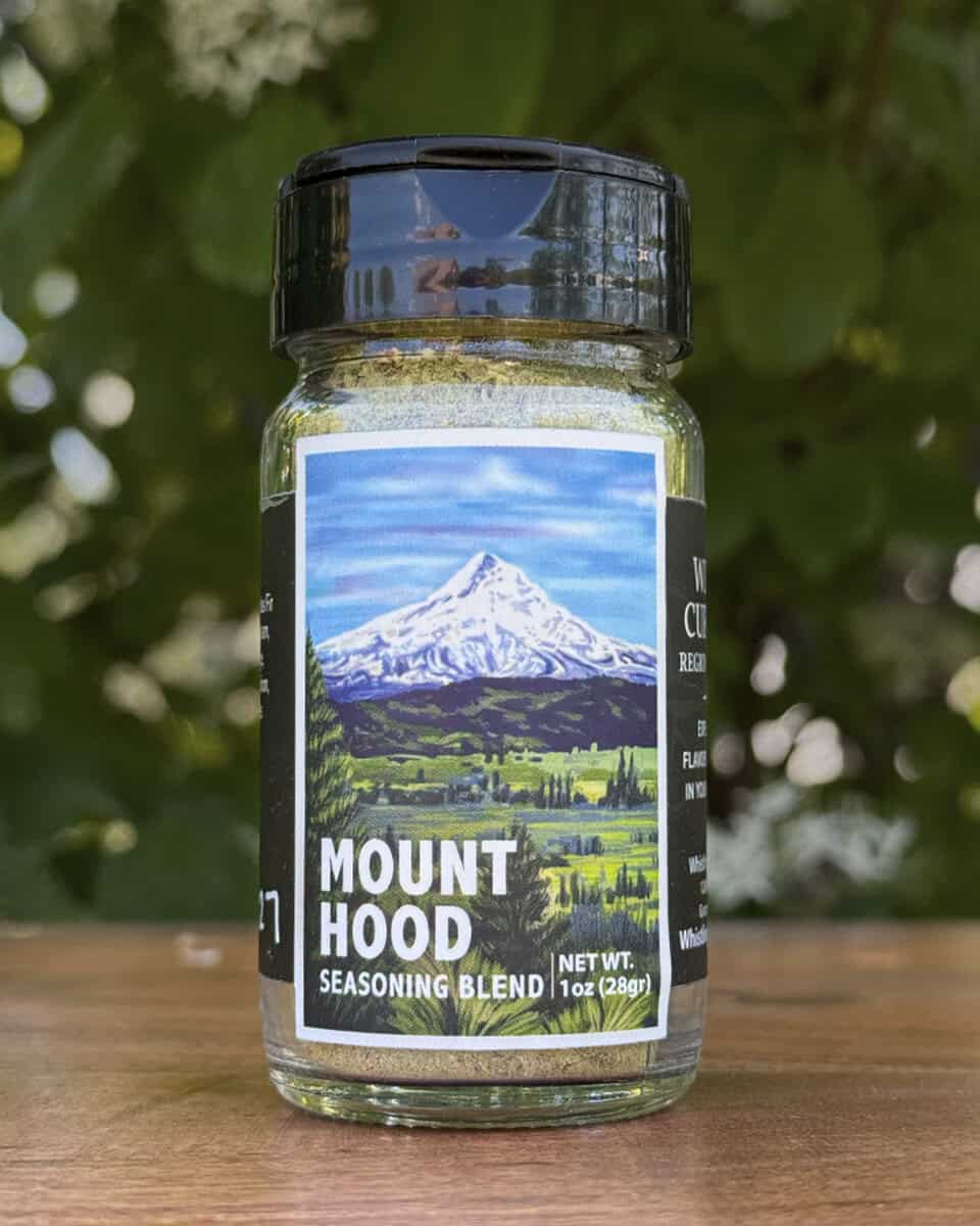 Glass jar of “Mount Hood” seasoning blend sitting on a wooden table outdoors with blurred green leaves and white flowers in the background. The label features an illustrated ice-capped mountain with a lush green foreground beneath a blue sky.