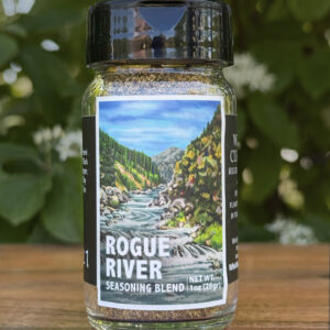 Glass jar of โRogue Riverโ seasoning blend sitting on a wooden table outdoors with blurred green leaves and white flowers in the background. The label features an illustrated mountain river flowing through a forest canyon beneath a blue sky.