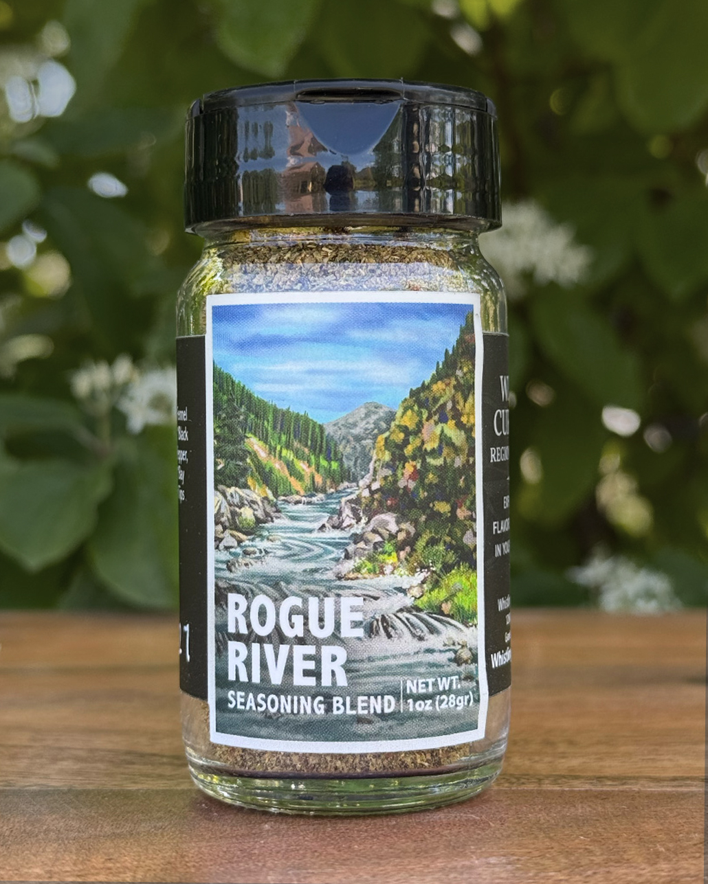 Glass jar of “Rogue River” seasoning blend sitting on a wooden table outdoors with blurred green leaves and white flowers in the background. The label features an illustrated mountain river flowing through a forest canyon beneath a blue sky.