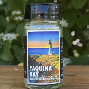 Glass jar of “Yaquina Bay” seasoning blend sitting on a wooden table outdoors with blurred green leaves and white flowers in the background. The label features an illustrated lighthouse on the coast beneath a sunset sky.
