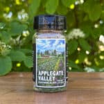 Glass jar of “Applegate Valley” seasoning blend sitting on a wooden table outdoors with blurred green leaves and white flowers in the background. The label features illustrated rows of crops on a farm beneath a blue sky.