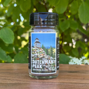 Glass jar of “Dutchman Peak” seasoning blend sitting on a wooden table outdoors with blurred green leaves and white flowers in the background. The label features an illustrated historic fire lookout on a mountaintop beneath a blue sky.