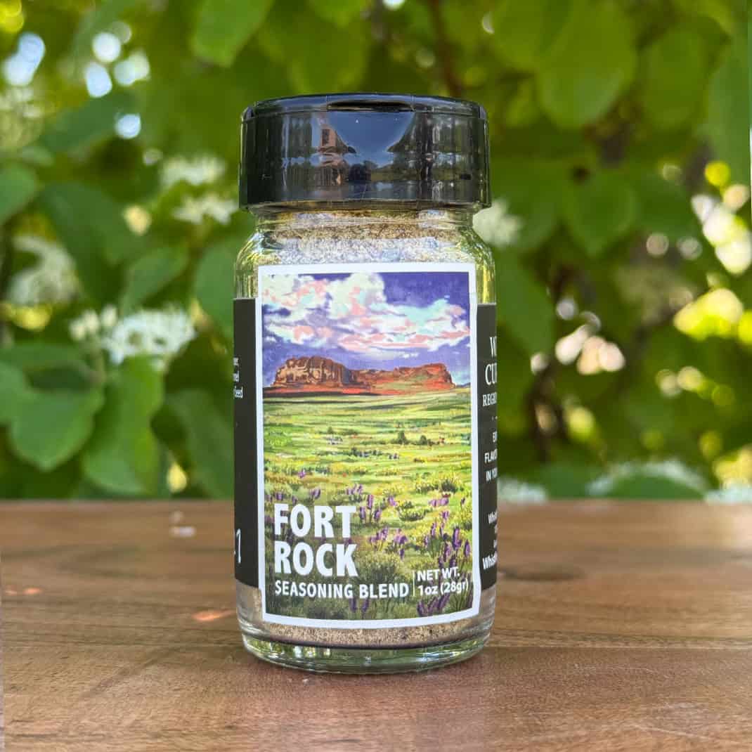 Glass jar of “Fort Rock” seasoning blend sitting on a wooden table outdoors with blurred green leaves and white flowers in the background. The label features an illustrated desert scape beneath a blue sky.
