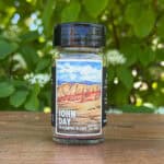 Glass jar of “John Day” seasoning blend sitting on a wooden table outdoors with blurred green leaves and white flowers in the background. The label features an illustrated desert scape beneath a blue sky.