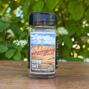 Glass jar of “John Day” seasoning blend sitting on a wooden table outdoors with blurred green leaves and white flowers in the background. The label features an illustrated desert scape beneath a blue sky.