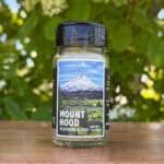 Glass jar of “Mount Hood” seasoning blend sitting on a wooden table outdoors with blurred green leaves and white flowers in the background. The label features an illustrated ice-capped mountain with a lush green foreground beneath a blue sky.