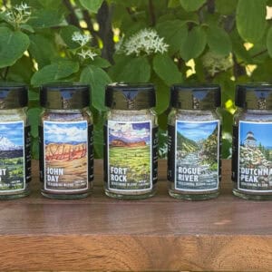 Seven small jars of Oregon-inspired seasoning blends lined up on a wooden table outdoors with leafy green plants and white flowers in the background. Each jar features illustrated landscape labels for “Applegate Valley,” “Mount Hood,” “John Day,” “Fort Rock,” “Rogue River,” “Dutchman Peak,” and “Yaquina Bay.”