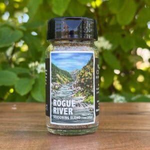 Glass jar of “Rogue River” seasoning blend sitting on a wooden table outdoors with blurred green leaves and white flowers in the background. The label features an illustrated mountain river flowing through a forest canyon beneath a blue sky.