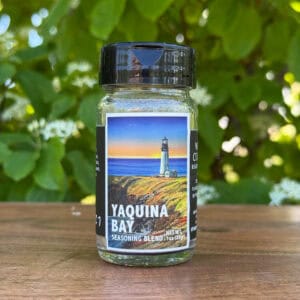 Glass jar of “Yaquina Bay” seasoning blend sitting on a wooden table outdoors with blurred green leaves and white flowers in the background. The label features an illustrated lighthouse on the coast beneath a sunset sky.