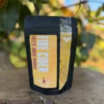 One resealable pouch of powdered food product sits on a wooden surface outdoors with blurred greenery in the background. The package shows a yellow label reading "Fire Cider Freeze-Dried Powder" from Whistling Duck Farm.