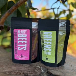 Two resealable pouches of powdered food products sit on a wooden surface outdoors with blurred greenery in the background. One package shows a pink label reading "Hearty Beets Fermented & Freeze-Dried" and the other shows a green powder labeled "Greens Freeze-Dried Powder & Things" from Whistling Duck Farm.